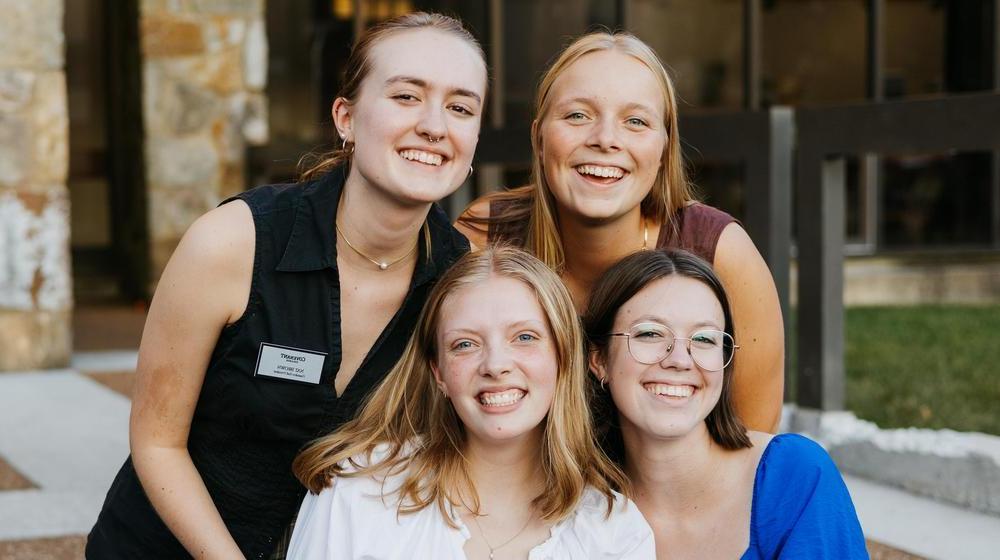 Four smiling young women posing together outdoors, with one wearing a name tag indicating student involvement.